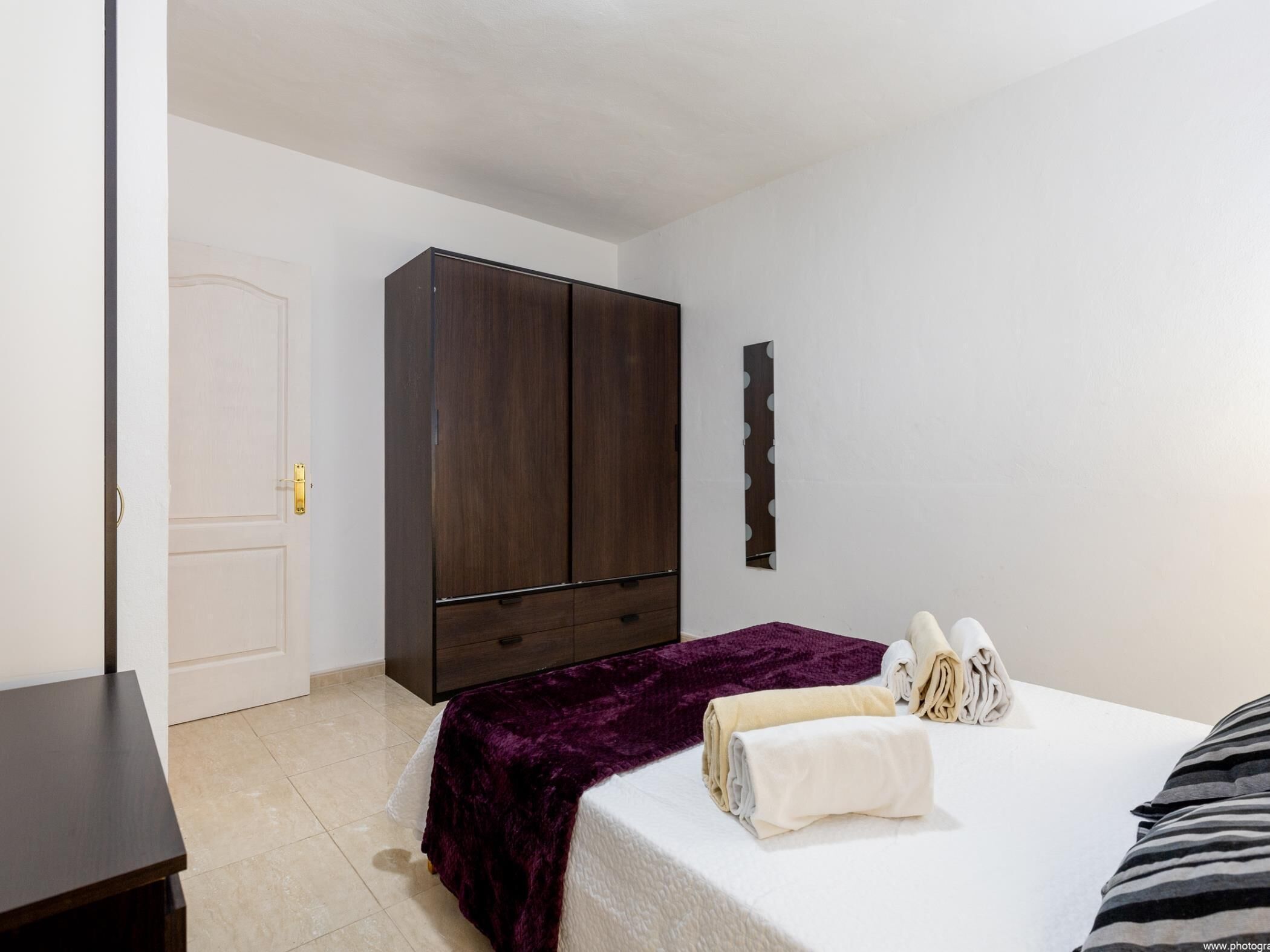 Quarto com guarda-roupa de madeira escura, cama com cobertor roxo e porta branca.