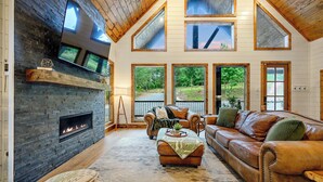 A spacious living room with a fireplace, leather sofas, and large windows offering views of the outdoors. - Broken Bow Home w/ Hot Tub, Game Room & EV Charger (Broken Bow)