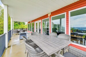 En overdækket terrasse med et bord og stole, store vinduer og udsigt til grønne områder.