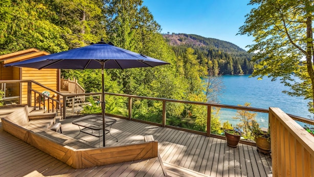 A wooden deck with a table and chairs, a blue umbrella, and a view of a lake surrounded by trees. - Reed s Cove Cabin in Hoodsport (Hoodsport)
