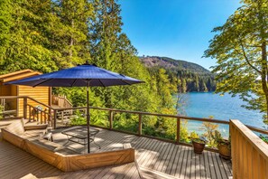 A wooden deck with a table and chairs, a blue umbrella, and a view of a lake surrounded by trees. - Reed s Cove Cabin in Hoodsport (Hoodsport)