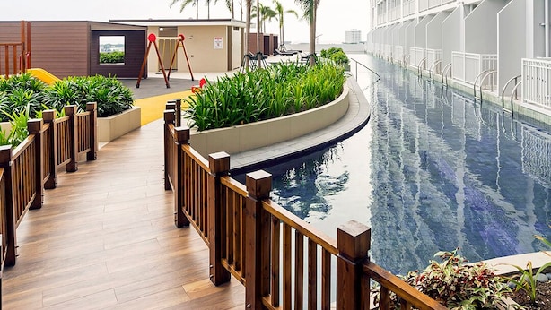 Zwembad met houten terras, omringd door planten en palmbomen, grenzend aan een modern gebouw met balkons.