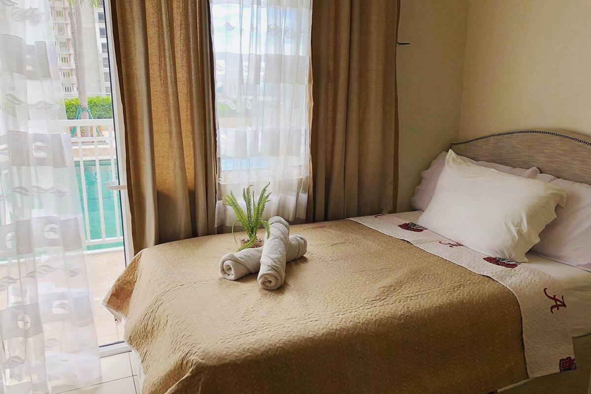 Ein Hotelzimmer mit Bett, Topfpflanze und Blick auf ein Gebäude sowie Grünfläche vor den Fenstern.