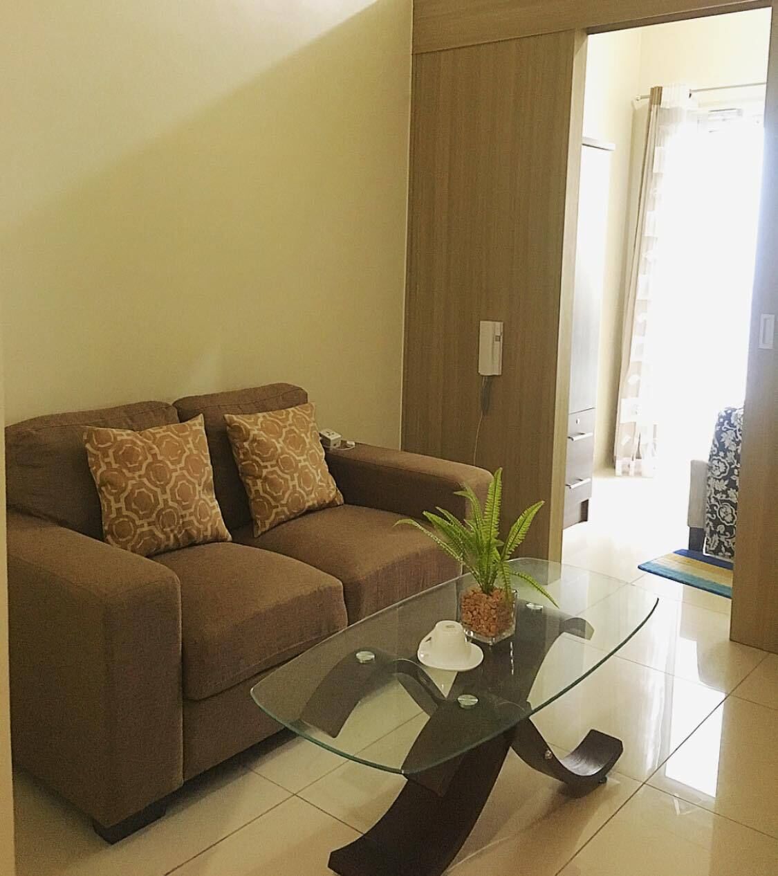 Een woonkamer met een bruine bank, twee gekleurde kussens, een glazen bijzettafeltje met een plant, en een moderne deur.