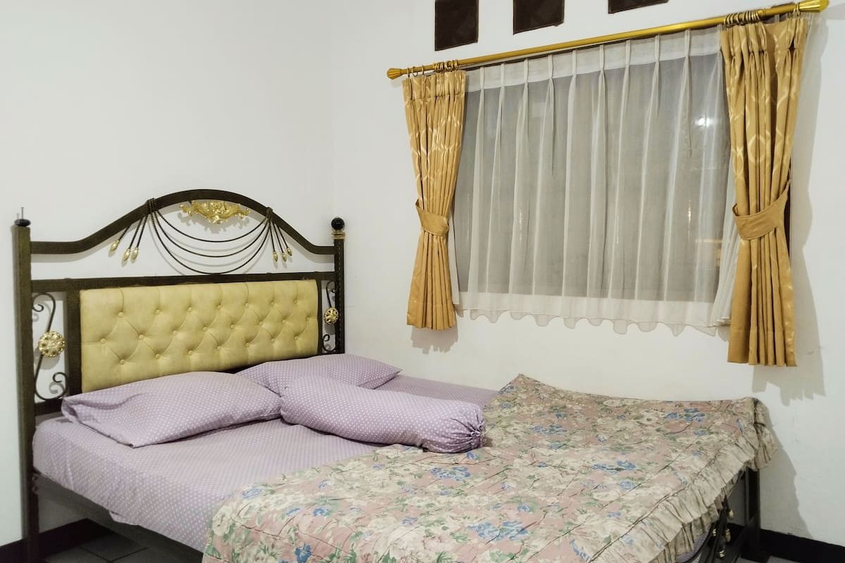Una cama con colcha estampada, cabecera capitonada y ventana con cortinas traslúcidas.