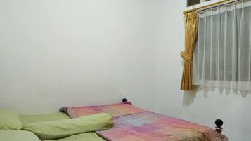 Una habitación pequeña con cama individual, pared blanca lisa, ventana con cortina translúcida y cama con manta colorida.
