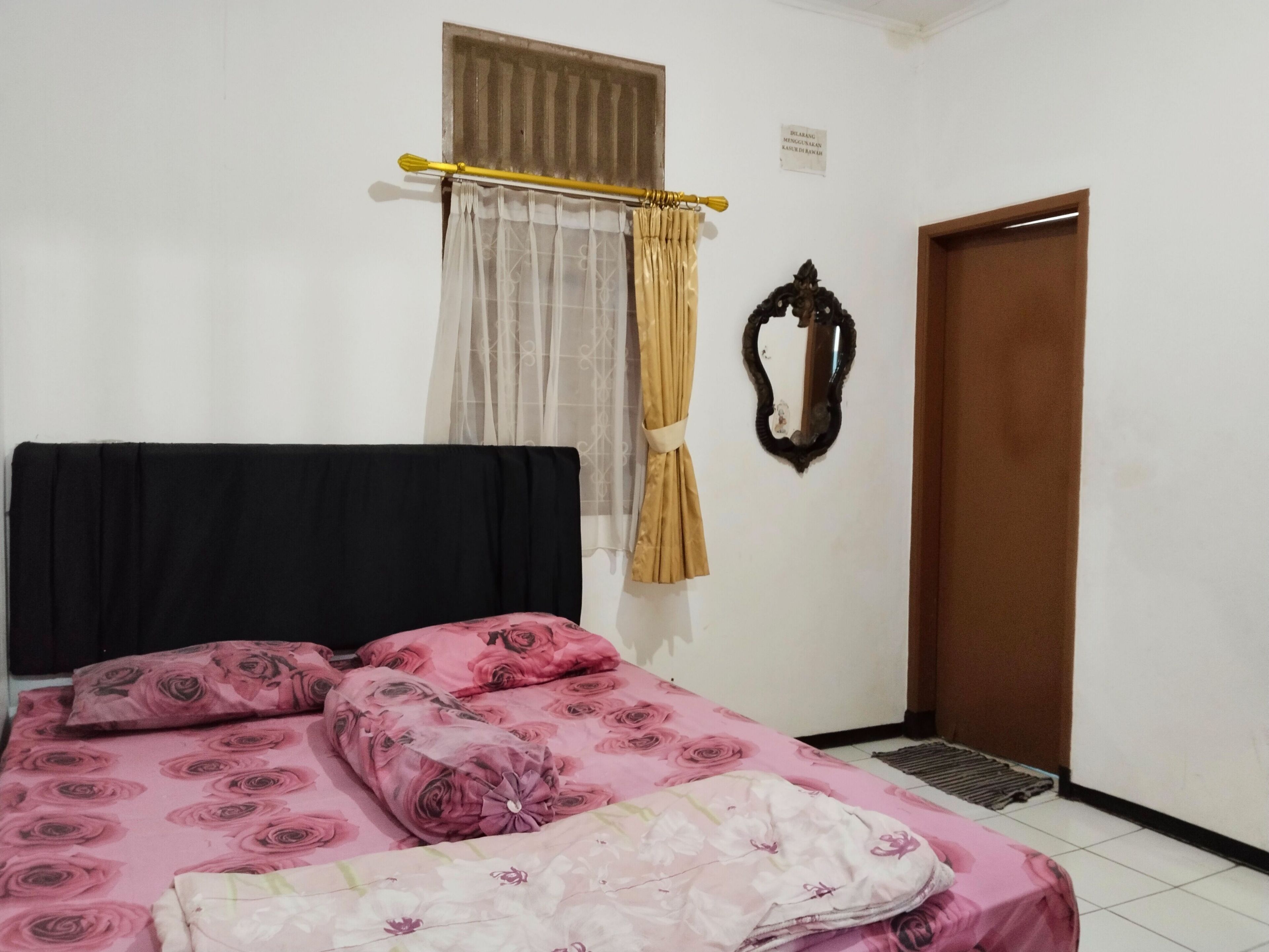 Un dormitori amb un llit, una finestra amb cortines, una porta i un mirall fixat a la paret.