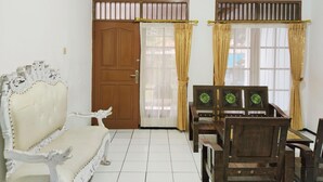 A room with a wooden door, a white upholstered sofa, a wooden dining table with chairs, and a window with curtains. - Villa Estria (Cisarua)
