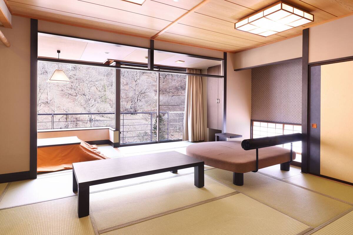 A traditional Japanese room with tatami flooring, a low table, a cushioned bench, and large windows offering a view of a snowy landscape.