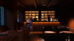 Un espace bar à la lumière tamisée, avec un barman, des étagères en bois où sont exposés divers objets et des coins salon.