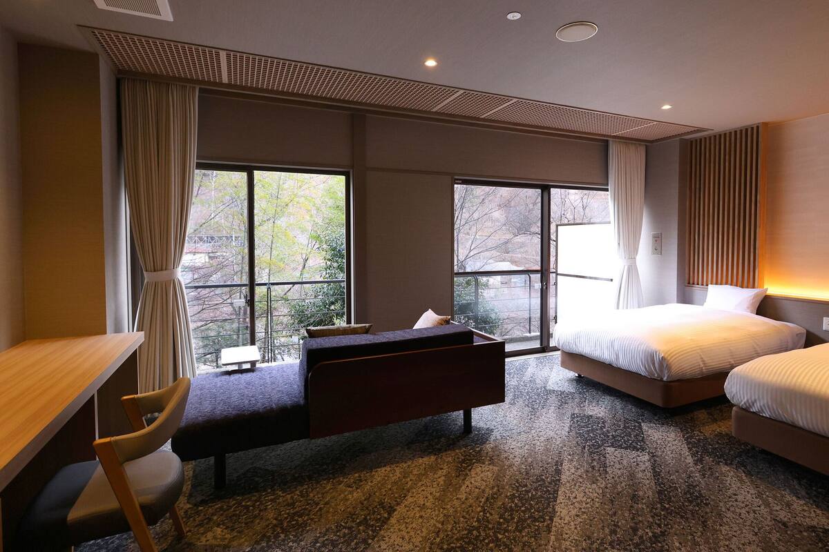A modern hotel room with a large window, a desk, and two beds.