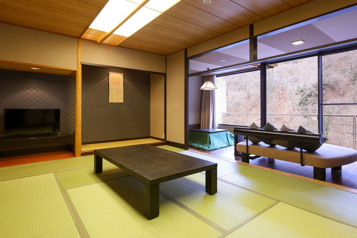 A tatami mat room with a low table, a television, and a view of trees through large windows.
