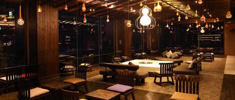 A cozy lounge area with wooden furniture, hanging pendant lights, and a fire pit.