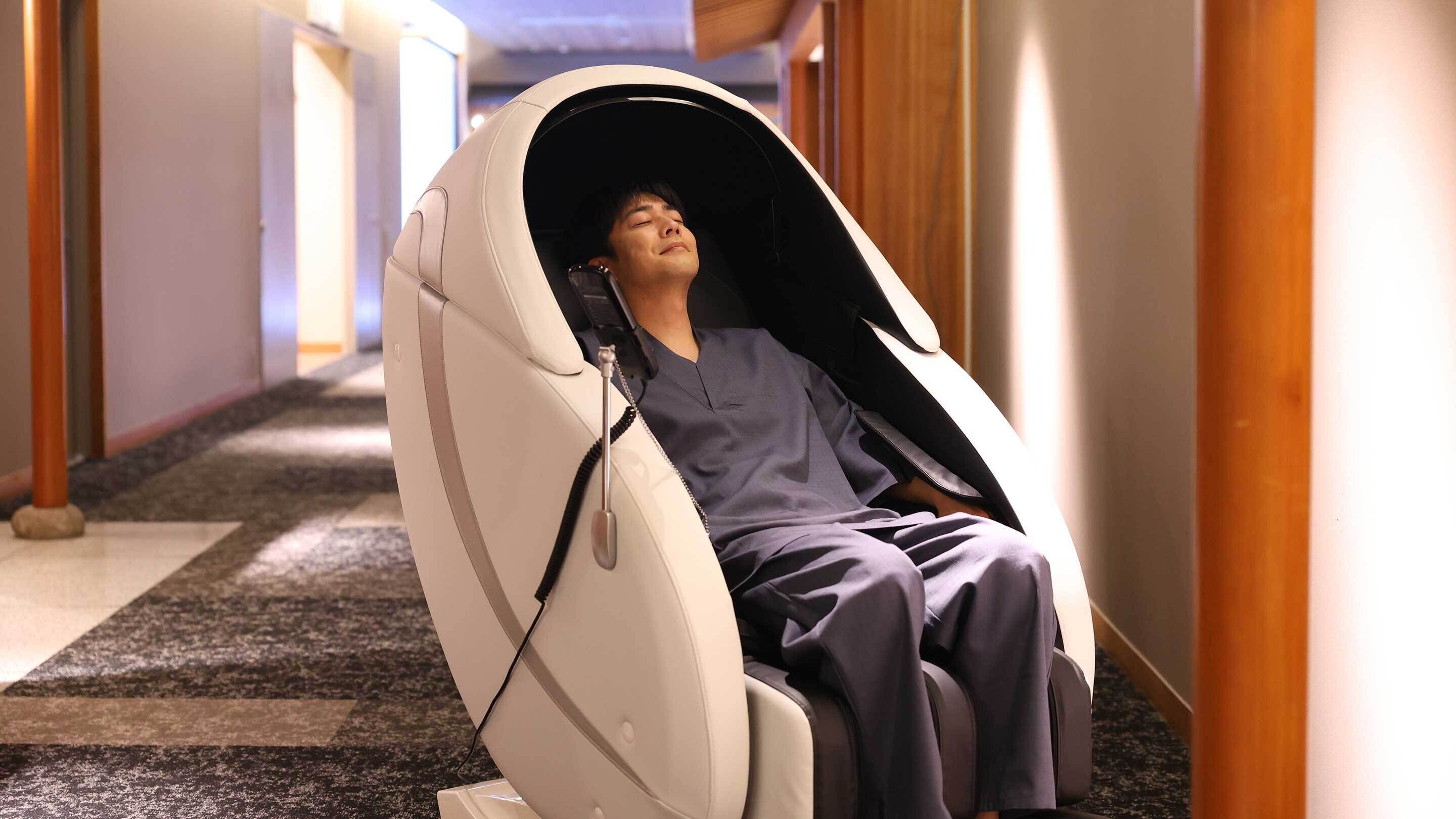 A person in a reclining chair with a white canopy, seated in a hallway with wooden flooring and walls.
