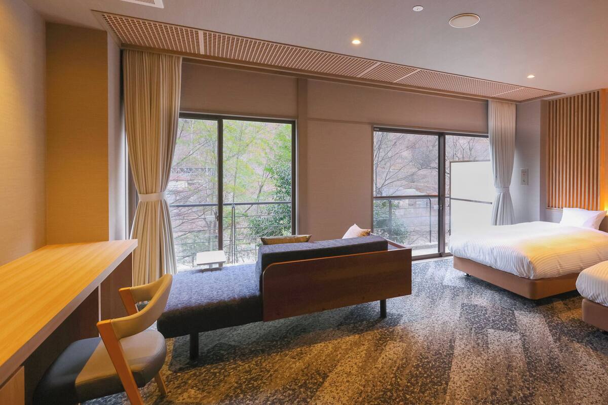 A hotel room with a desk, two beds, a chair, and large windows.
