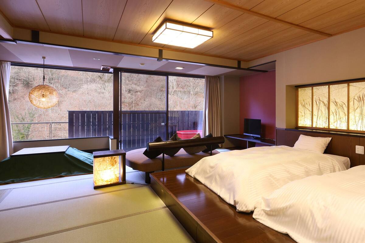 A hotel room with two beds, a futon, a TV, and a large window with a view of trees.