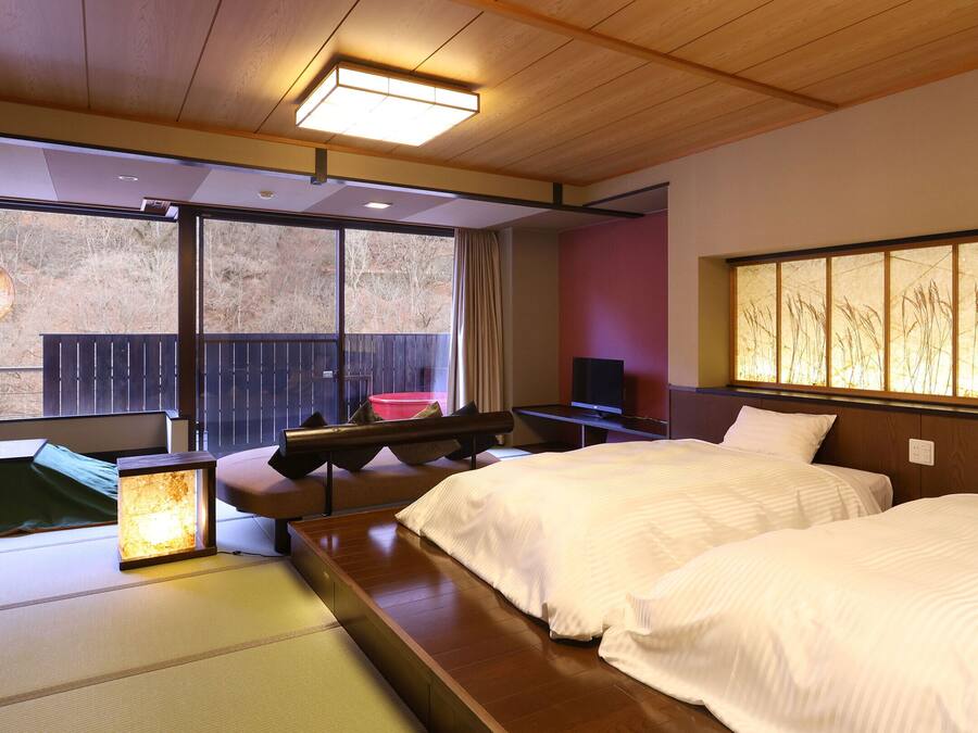 Quarto de hotel com duas camas, um futon, uma televisão e uma janela grande com vista para árvores.