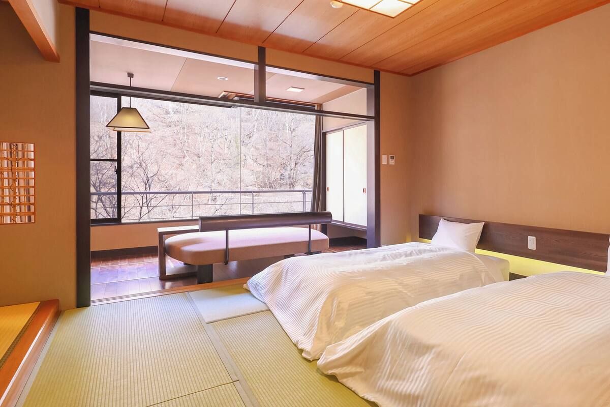 A hotel room with tatami flooring, a wooden ceiling, a bench by the window, and two beds with white bedding.