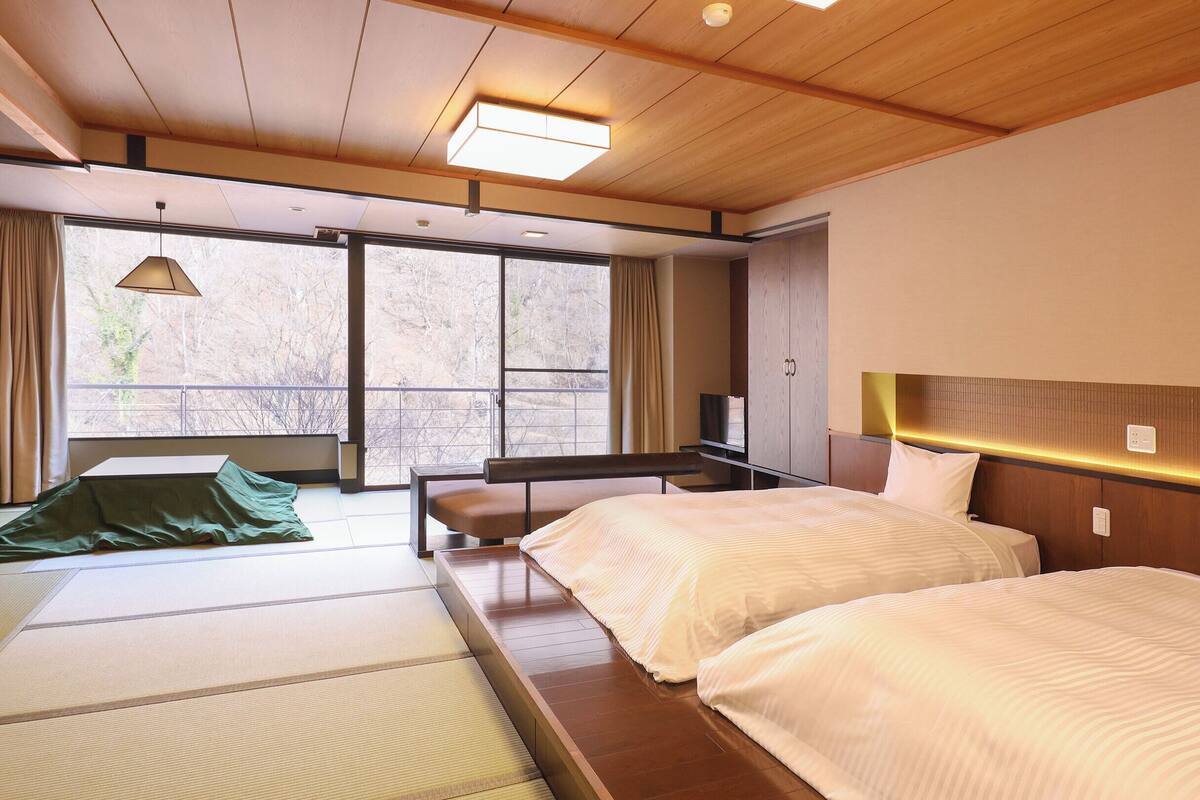 A hotel room with two beds, a sofa, a TV, and a large window with a view of trees.