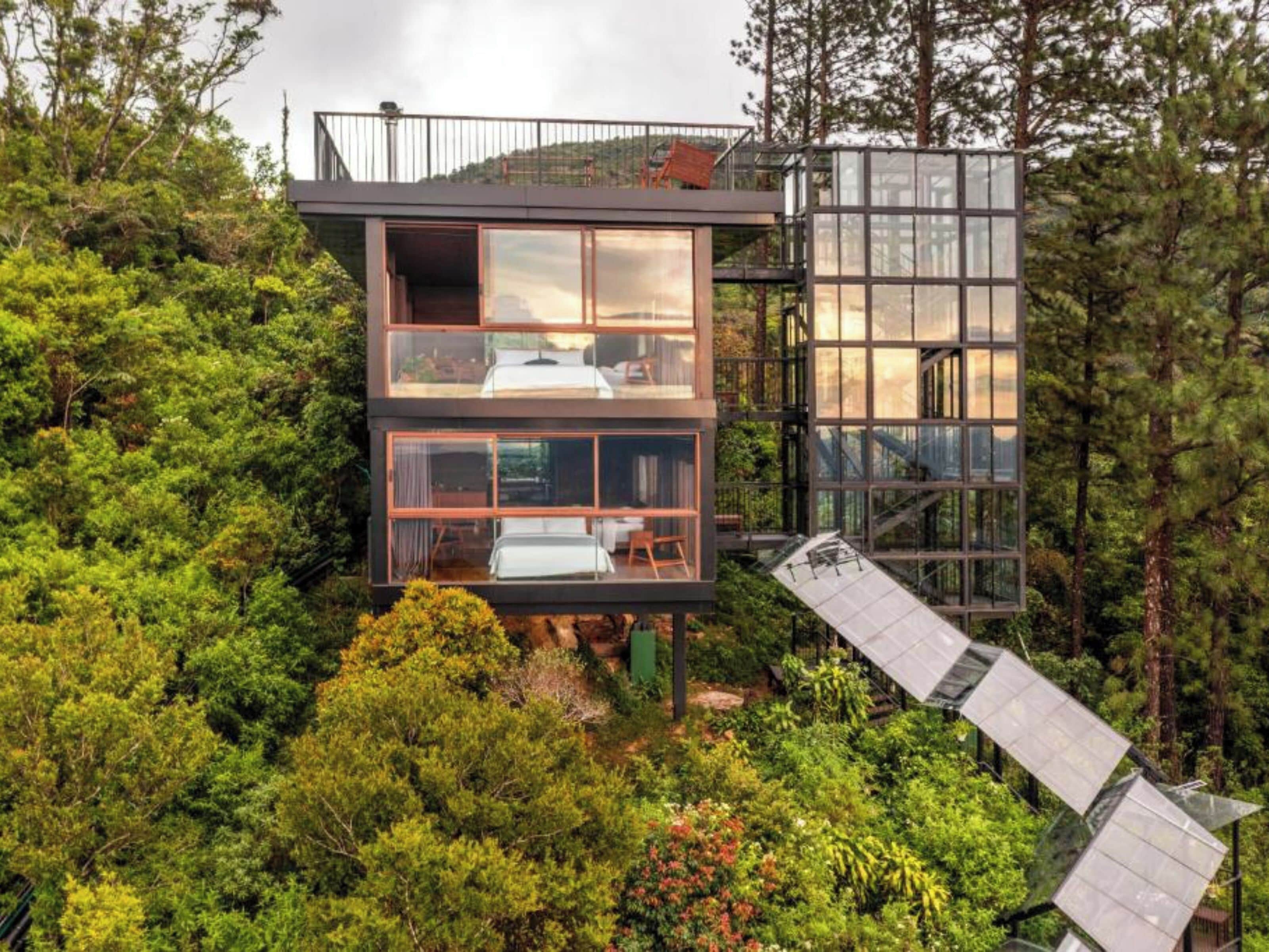 A modern, multi-level cabin nestled in a forest. It features large glass windows, a balcony, and a unique sloped roof design.