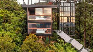 A modern, multi-level cabin nestled in a forest. It features large glass windows, a balcony, and a unique sloped roof design.