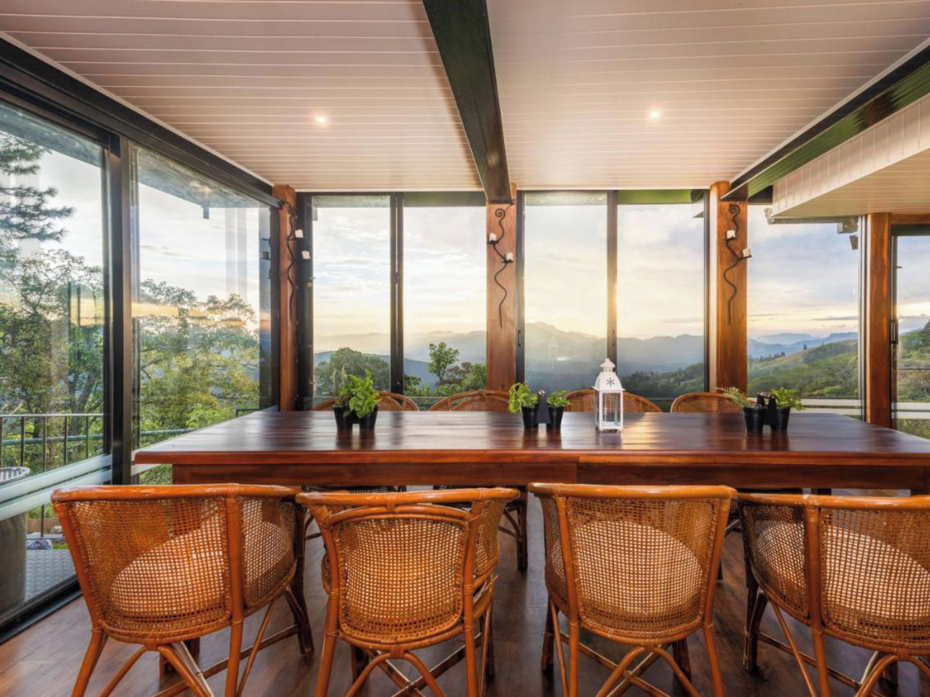 Un comedor con una mesa de madera y sillas de mimbre, grandes ventanales que ofrecen vistas a un paisaje montañoso.