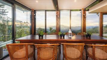 Un comedor con una mesa de madera y sillas de mimbre, grandes ventanales que ofrecen vistas a un paisaje montañoso.