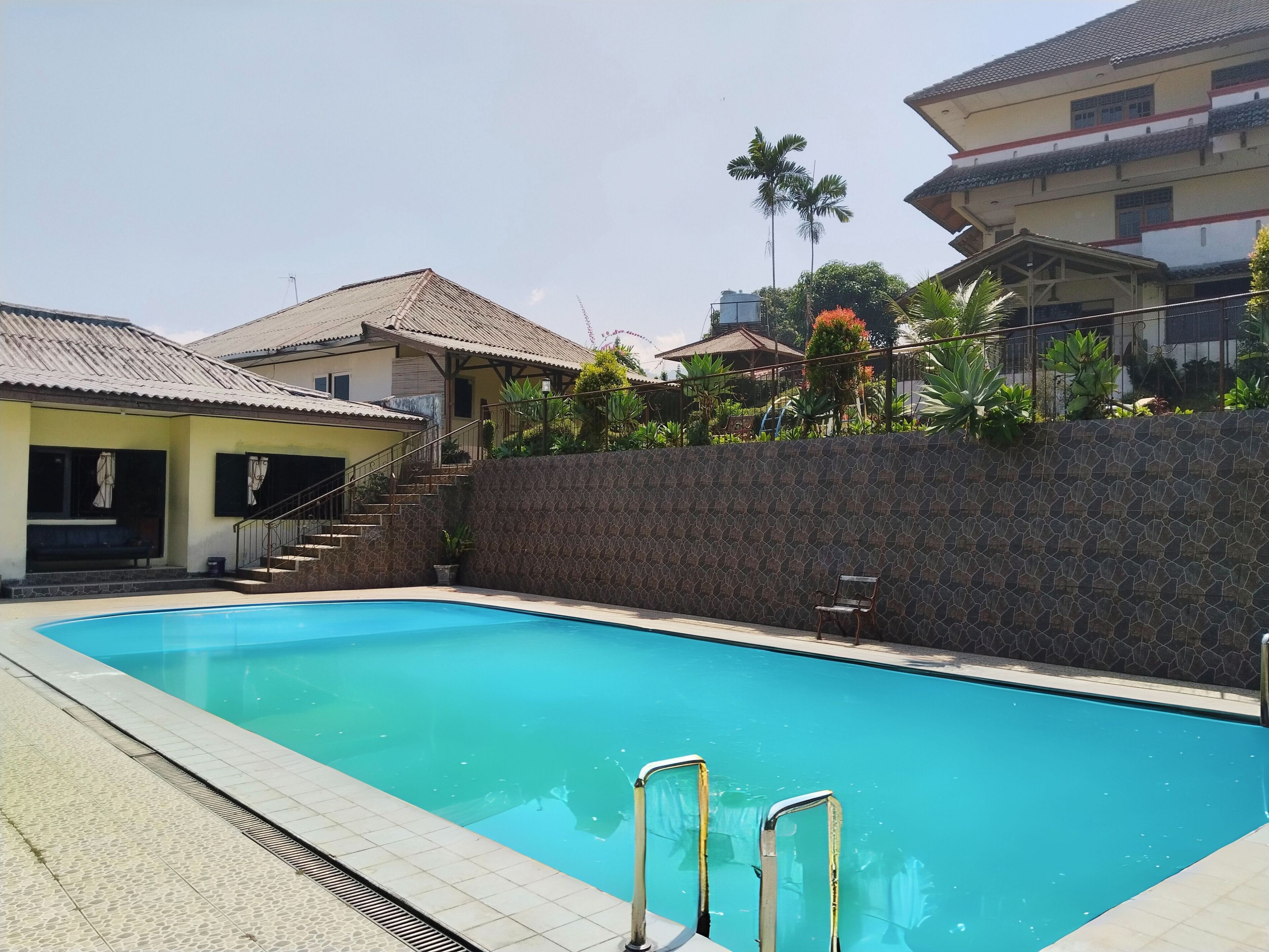 A swimming pool with steps leading into it, surrounded by a stone wall and a building with a tiled roof.
