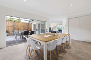 A modern dining area with a wooden table and white chairs, a glass door leading to an outdoor patio, and a view of a blue sofa and a clock on the wall.