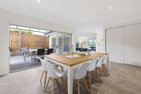 A modern dining area with a wooden table and white chairs, a glass door leading to an outdoor patio, and a view of a blue sofa and a clock on the wall.
