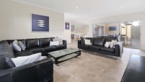 A modern living room with two black leather sofas, a glass coffee table, and a dining area in the background.