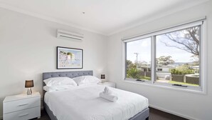 A bedroom with a large bed, two bedside tables, a wall-mounted air conditioner, a framed picture, and a window with a view of the outside.