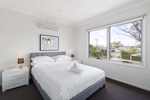 Une chambre à coucher avec un grand lit, deux tables de chevet, un climatiseur fixé au mur, un tableau encadré et une fenêtre donnant sur l’extérieur.