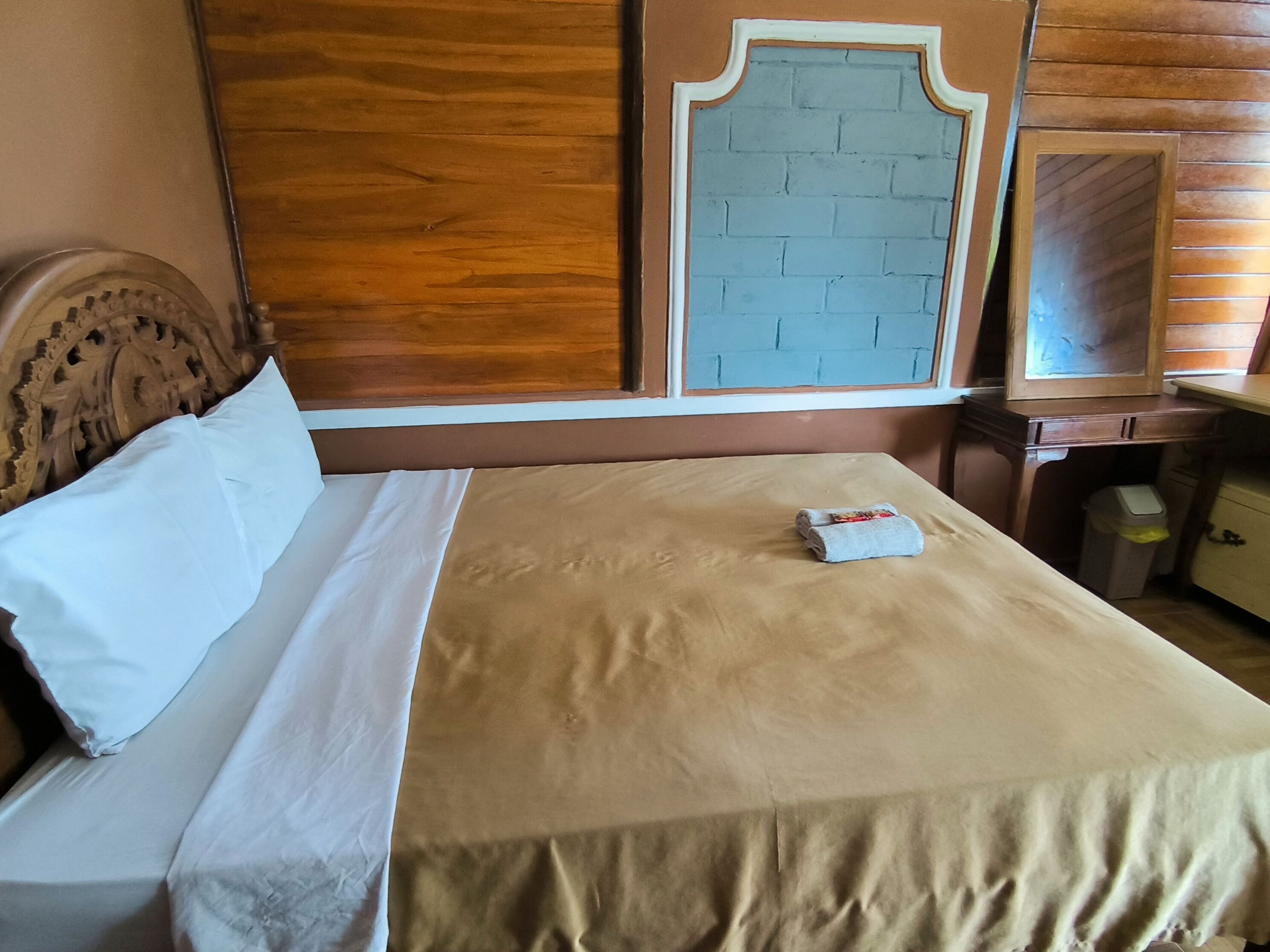 A neatly made bed with a beige bedspread and white pillows, a wooden headboard, and a wall with a brick-patterned design.