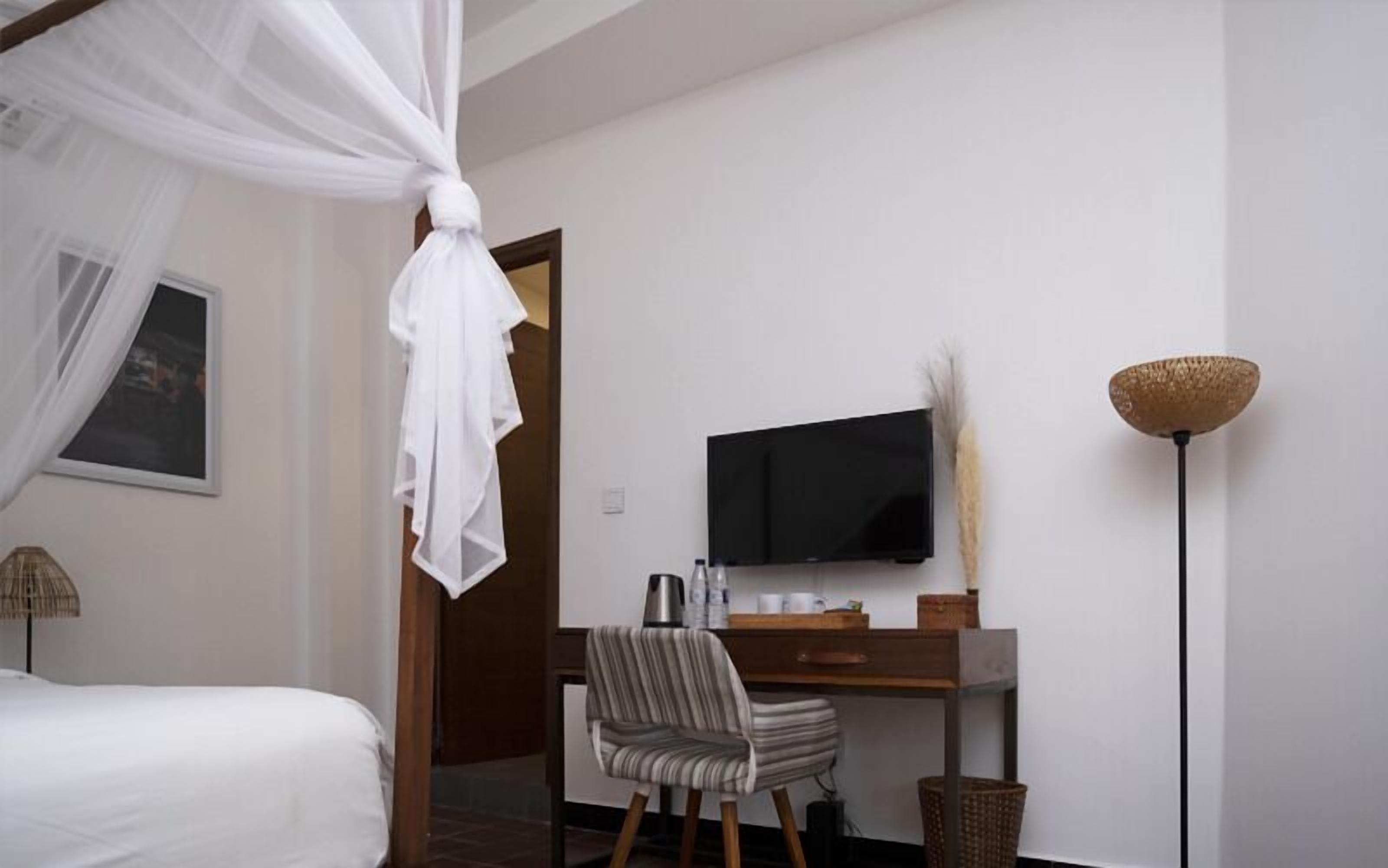 Un dormitorio con una cama, un escritorio con televisor, una silla, una mesita de noche y una lámpara de pie.