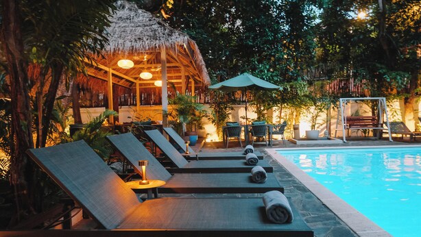 A poolside area with lounge chairs, a thatched-roof structure, and a small table with a lamp. - Jungle Addition (Phnom Penh)