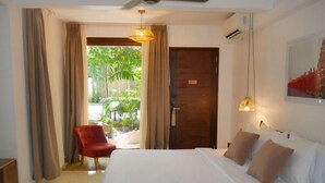 A hotel room with a large bed, a red armchair, and a view of a garden through a glass door. - Jungle Addition (Phnom Penh)