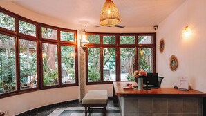 A reception area with a wooden bench, a desk with a computer, and a large window offering a view of greenery. - Jungle Addition (Phnom Penh)