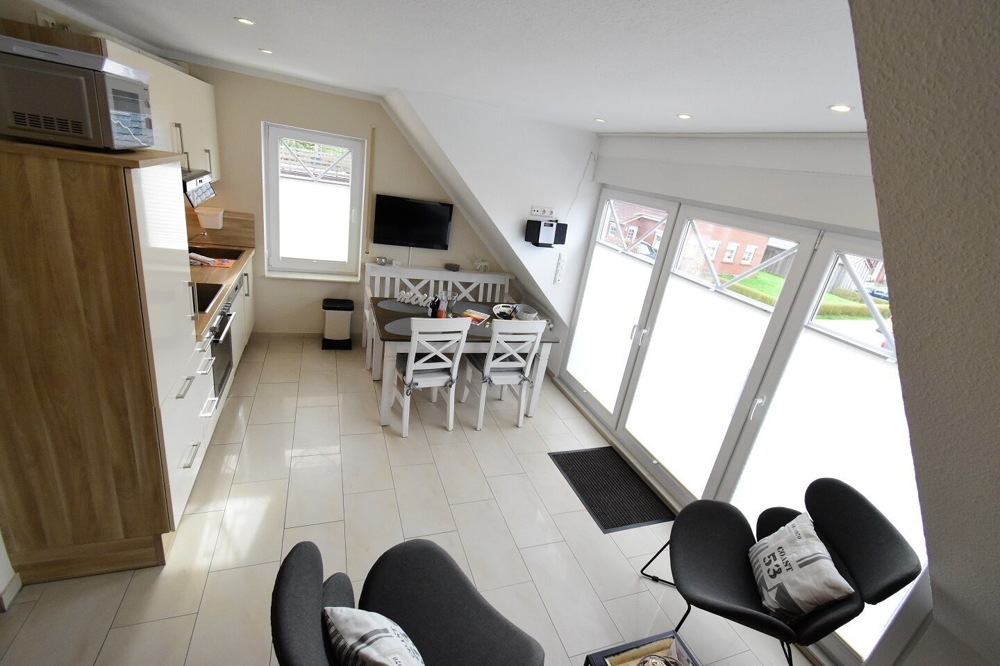 A modern kitchen with white cabinets and a built-in microwave, a dining area with a white table and chairs, and large windows offering a view of the outside.