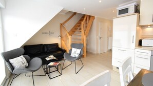 Un appartement moderne doté d’un salon comprenant un canapé en cuir noir, deux fauteuils, une table basse et une table à manger. Un escalier en bois mène à une chambre.