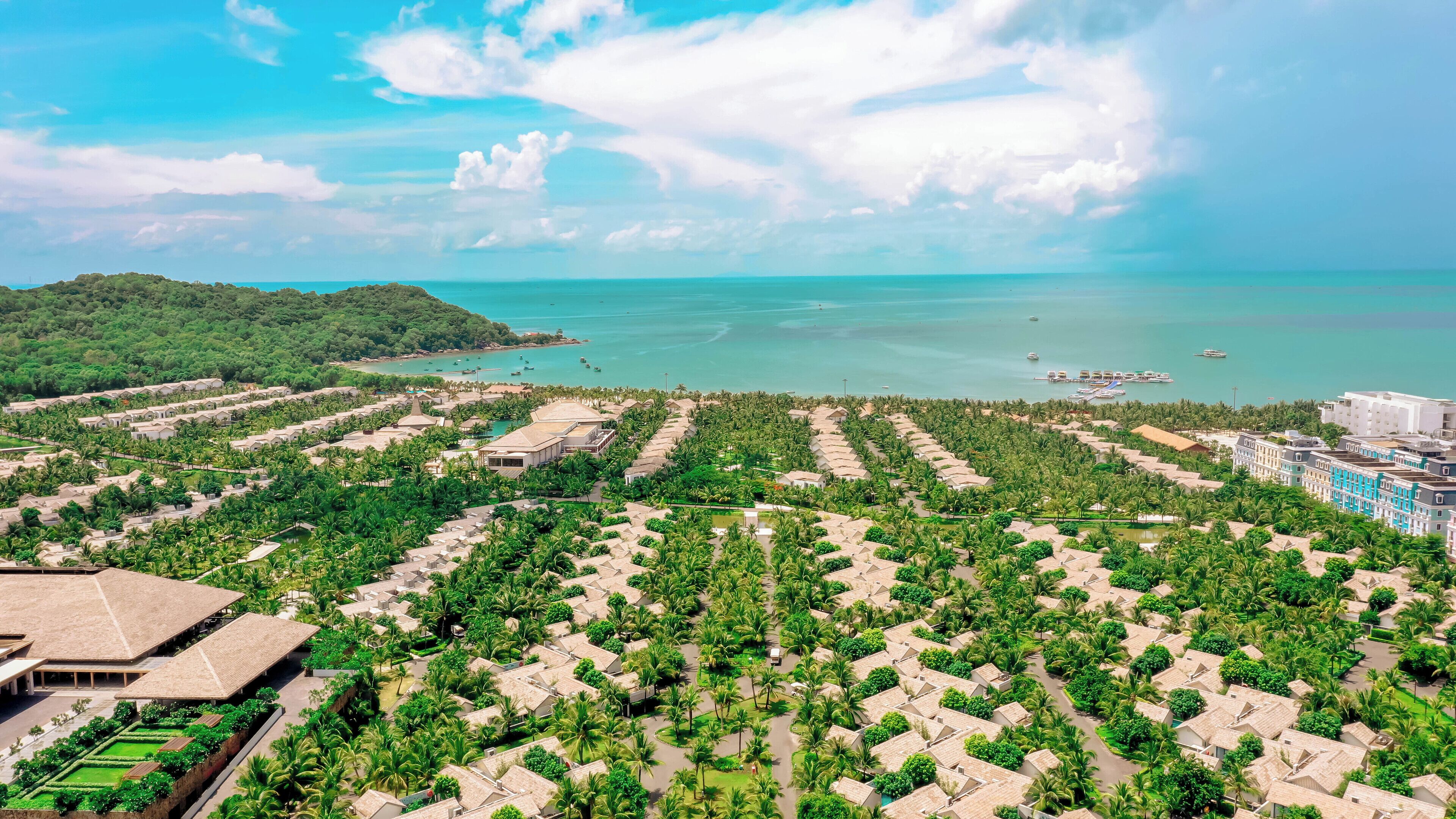 Vista aérea de un resort con varias cabañas de estilo bungalow dispuestas en filas, rodeado de vegetación exuberante, y con vista al mar y a los barcos en el fondo.