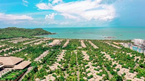 An aerial view of a resort with multiple bungalow-style accommodations arranged in rows, surrounded by lush greenery, with a view of the sea and boats in the background. - HL Melody Boutique Hotel (Phu Quoc)