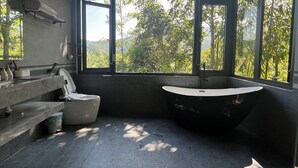 Une salle de bain moderne avec une baignoire sur pieds, des toilettes et un lavabo. De grandes fenêtres offrent une vue sur la verdure environnante.