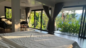 Une chambre moderne avec un grand lit, un canapé et une vue imprenable sur une végétation luxuriante et des montagnes, grâce à de grandes fenêtres.