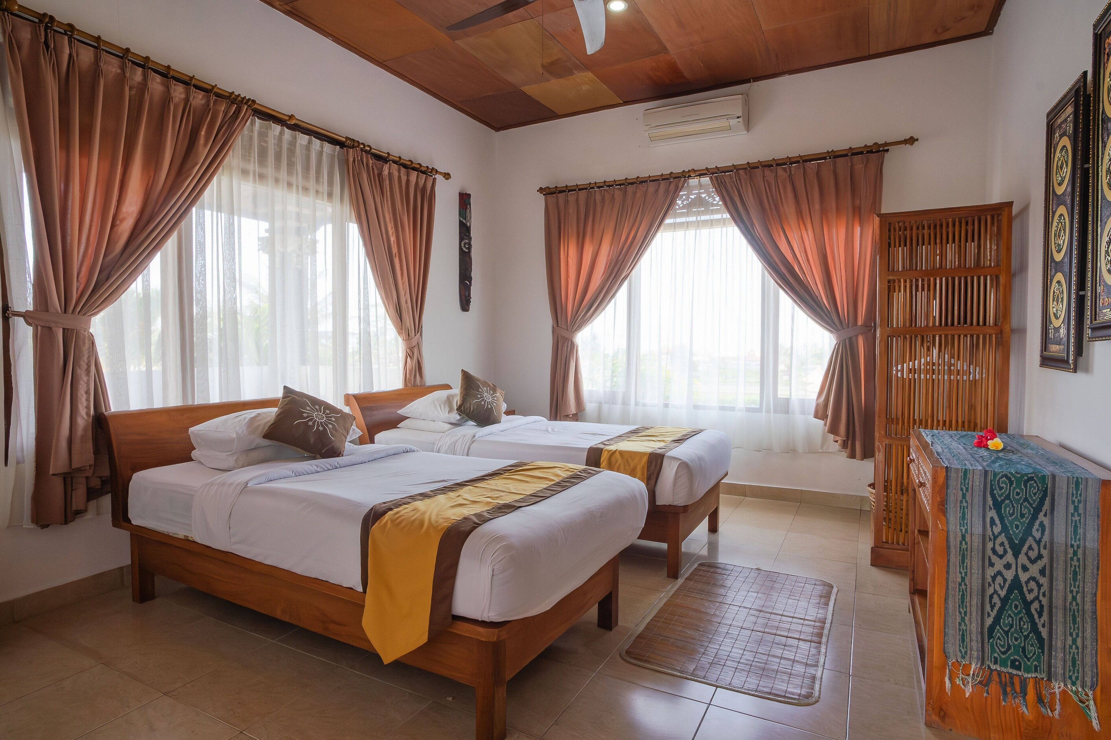 A hotel room with two wooden beds, beige curtains, a wooden headboard, a small table with a patterned cloth, and a wall-mounted air conditioner.