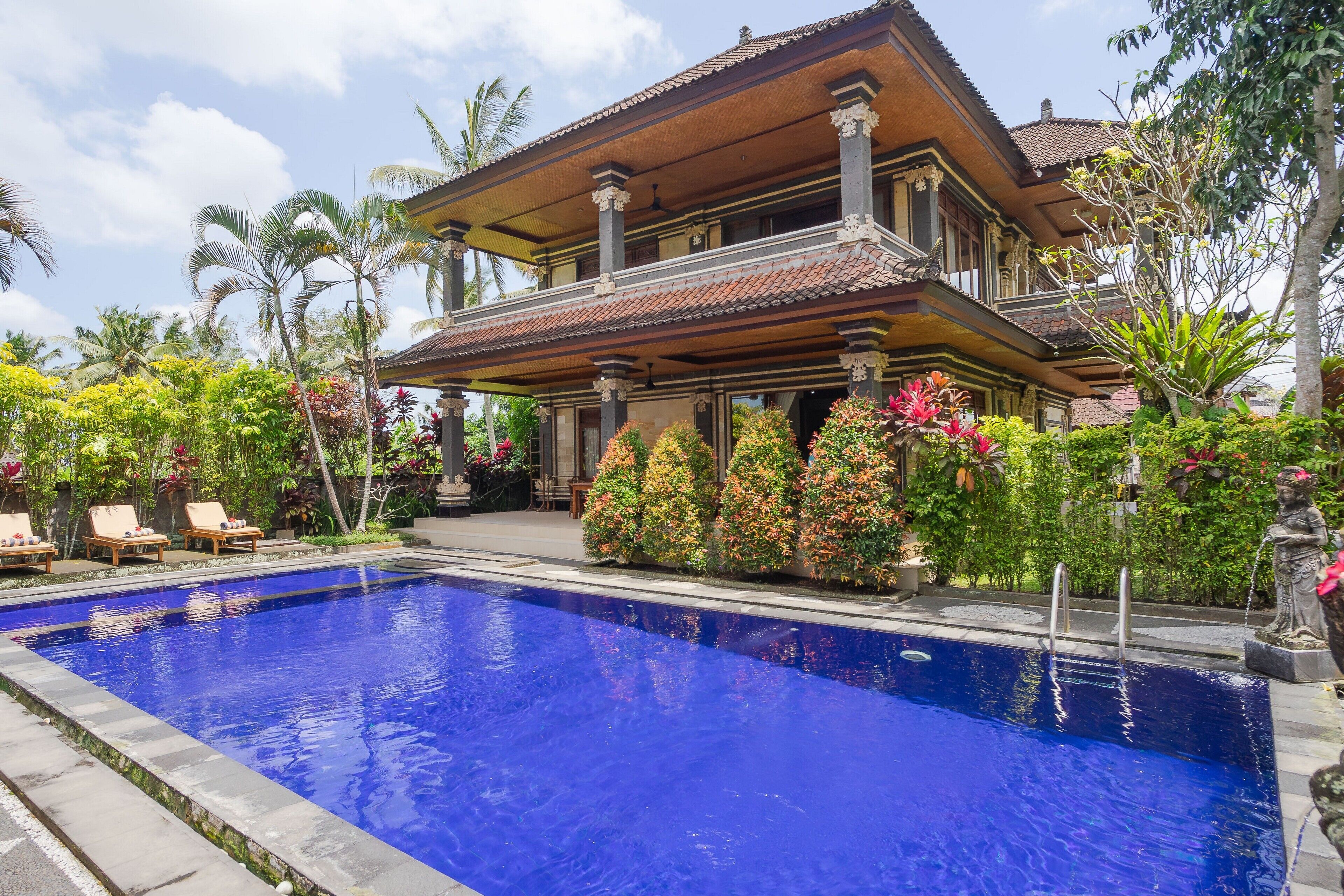 A large swimming pool with a wooden deck and lounge chairs, surrounded by lush tropical plants and a traditional house with a tiled roof.