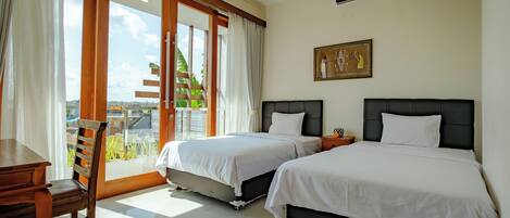 A hotel room with two beds, a wooden desk, and a view of greenery through a sliding glass door.