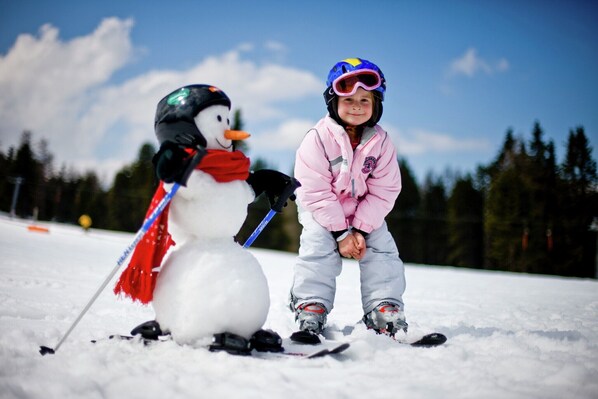 A child standing next to a snowman with a red scarf and a blue scarf, wearing a pink jacket and a helmet, in a snowy outdoor setting.