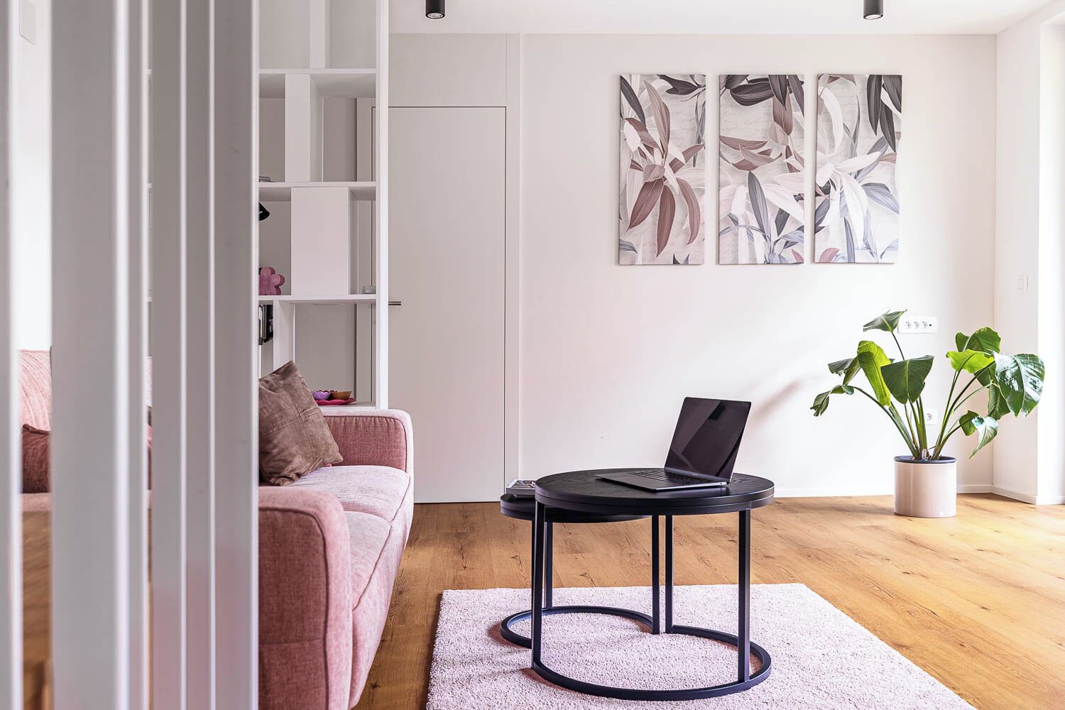 Ein modernes Wohnzimmer mit einem rosafarbenen Sofa, einem runden schwarzen Tisch mit einem Laptop, einer Topfpflanze und abstrakter Wandkunst.