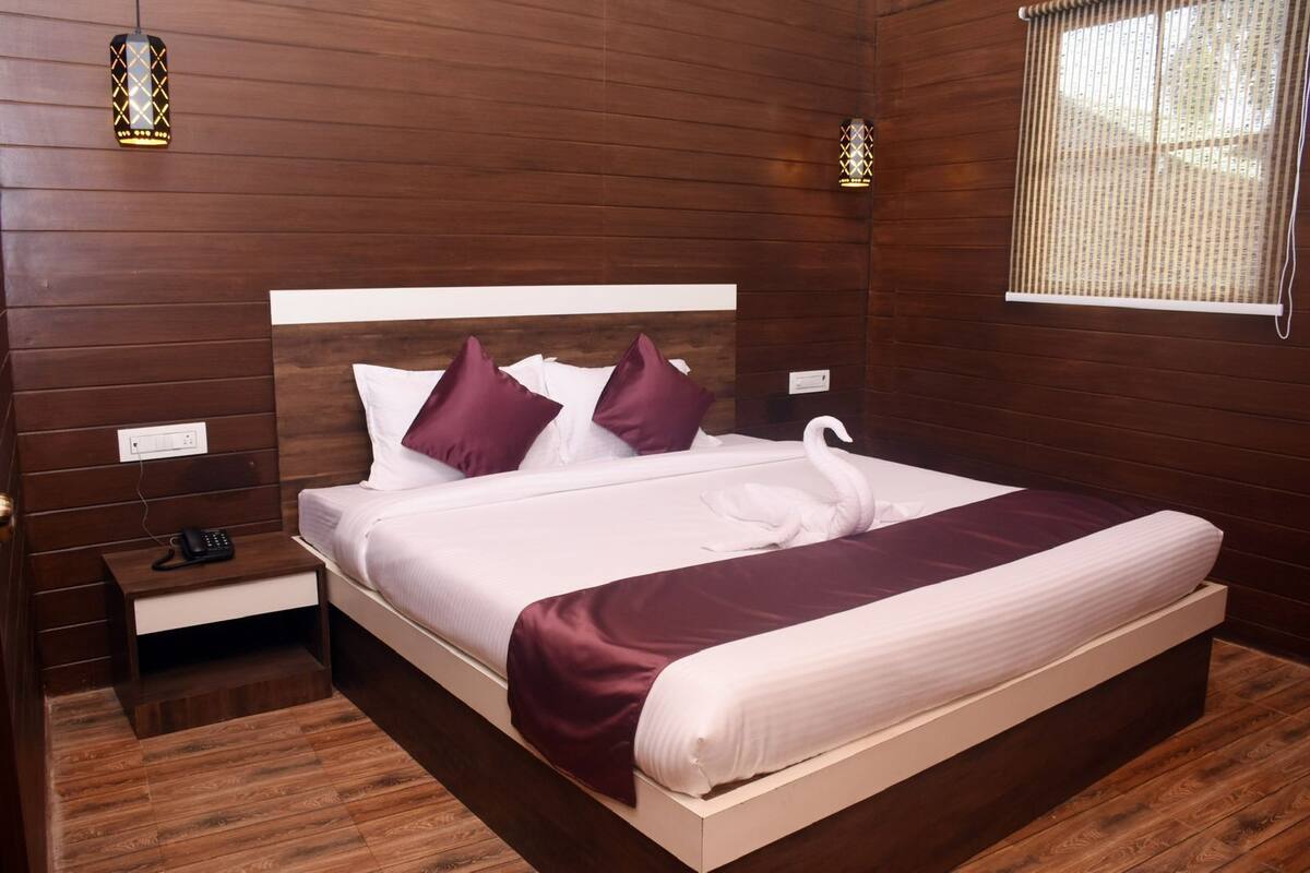 A neatly made bed with a white headboard, two pillows, and a decorative swan. There is a nightstand with a phone and a lamp. The room has wooden walls and a window with blinds.